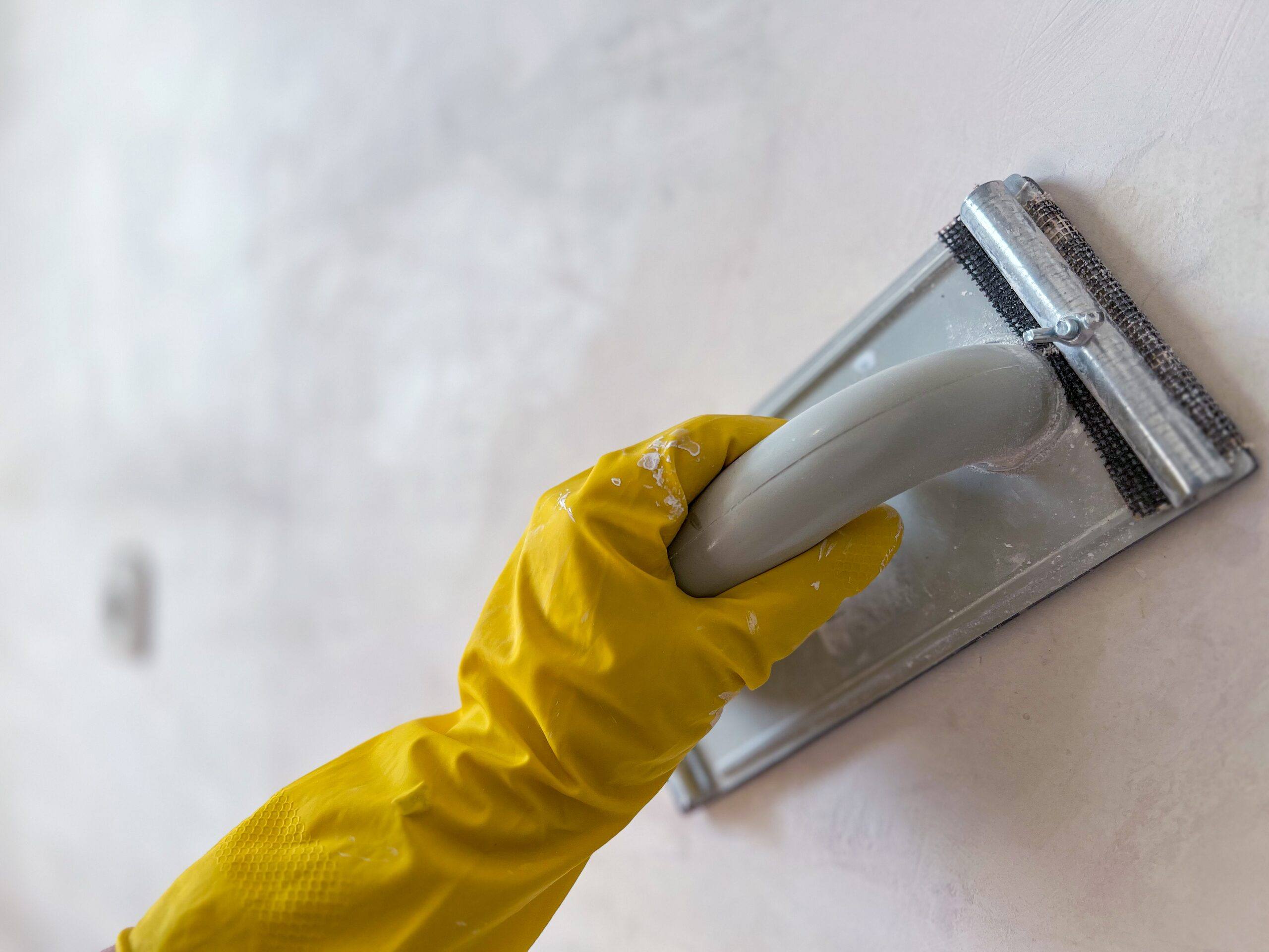 Mano con guante amarillo lijando una pared enmasillada con un taco lijador