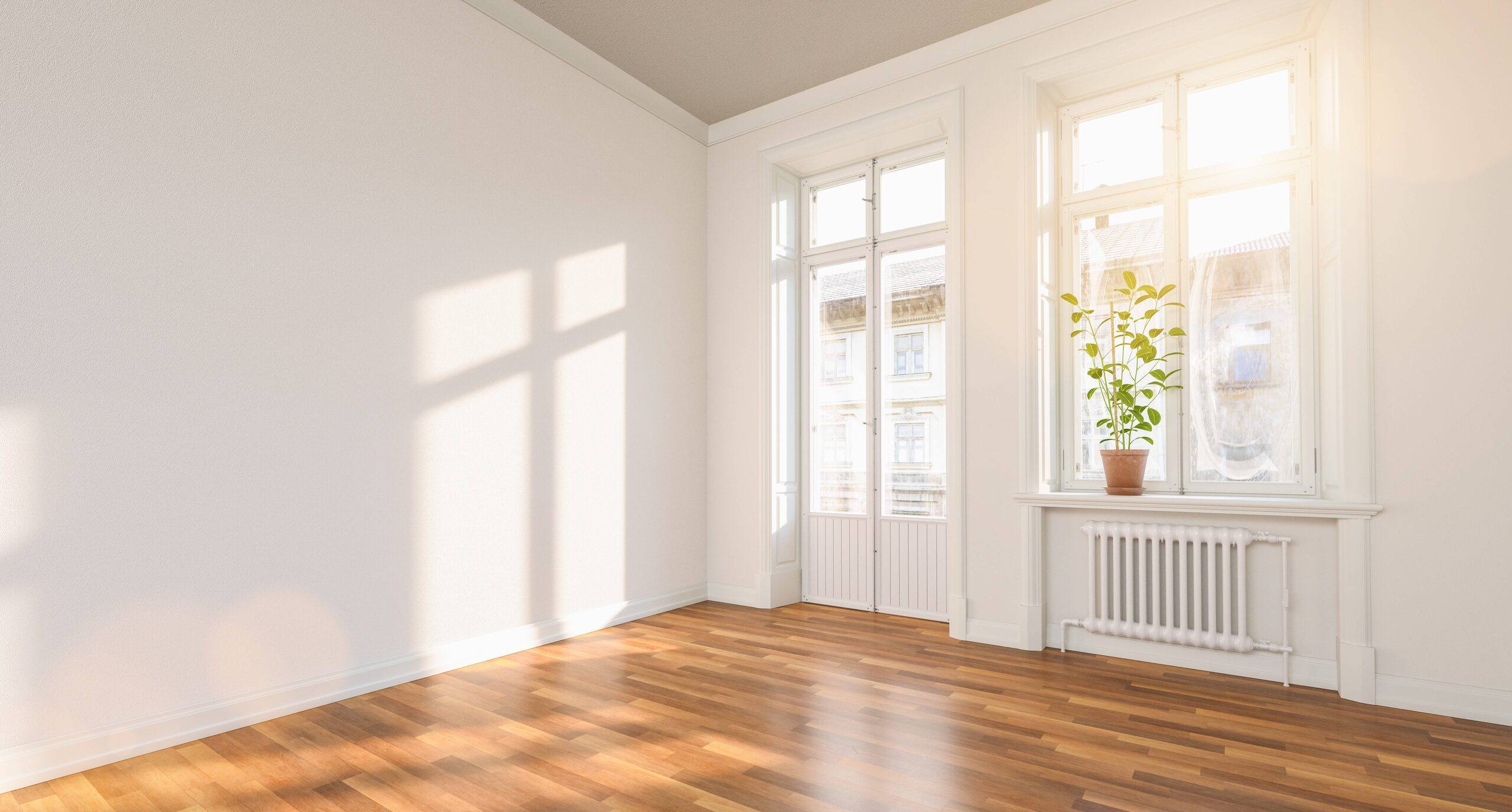 Salón vacío recién pintado en blanco roto con balconera, ventana con planta y luz de sol cálida sobre el suelo de madera